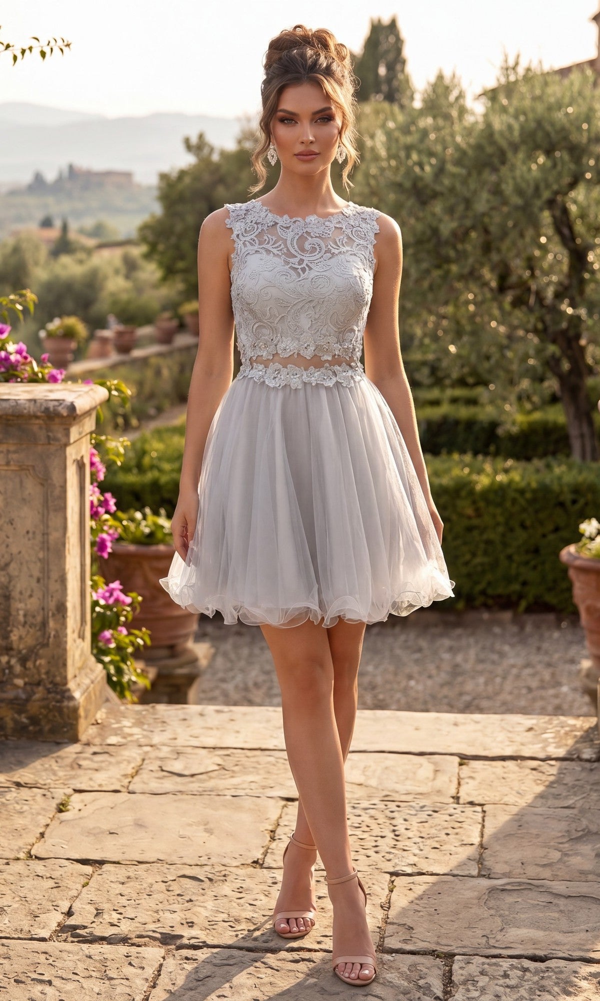 A woman stands outdoors on a stone patio surrounded by greenery and flowers, wearing the Short Homecoming Dress: Chicas C9350 in silver. This sleeveless dress features an illusion lace bodice and an elegant a-line tulle skirt, with a distant landscape visible in the background.